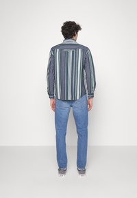Camisa a rayas en azul marino, verde y blanco, con cuello abotonado y mangas largas, combinada con jeans de un azul claro y zapatillas grises.