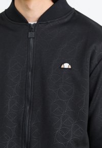 Black zip-up jacket with subtle embossed logo pattern, ribbed collar, and a small contrasting logo badge on the left chest area.