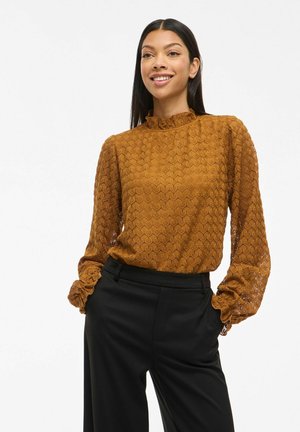 Blouse marron à motifs avec un col haut à volants et des manches amples, associée à un pantalon noir ajusté. Tissu texturé et léger.