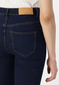 Jean en denim bleu foncé avec poche arrière visible et étiquette de marque en cuir marron "Noisy May" sur la ceinture, associé à un haut blanc texturé.