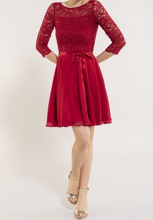 Frau in knielangem rotem Kleid mit Spitzenoberteil, dreiviertel Ärmeln, Satin-Taillenband und goldfarbenen offenen Zehabsätzen vor hellem Hintergrund.