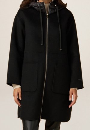 Woman wearing a black knee-length coat with front zipper, two large pockets, long sleeves, and a hood with drawstrings.