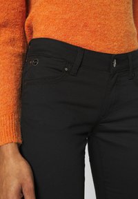 Vaqueros negros de corte slim, con costuras en contraste, herrajes de metal y cintura alta, combinados con un suéter naranja.