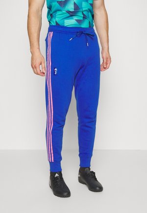 Homme portant un pantalon de survêtement bleu Adidas Juventus avec des rayures roses et des chaussures de football Adidas noires, debout devant un fond blanc.