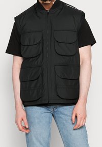 Musta hihaton liivi, joka on valmistettu teksturoidusta kankaasta, jossa on etuvetoketju, kuusi isoa taskua ja resorikaulus. Päällä mustan paidan päällä.