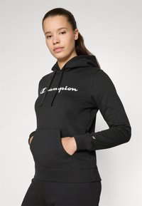 Svart hettegenser med frontlomme og snorhette. Har hvit "Champion" logo og ribbestrikkede mansjetter. Laget av myk, teksturert stoff.