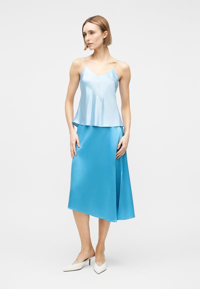 Γυναίκα που στέκεται φορώντας ένα γαλάζιο σατέν καμισόλι και ταιριαστό midi φούστα με λευκά μυτερά τακούνια σε απλό φόντο.