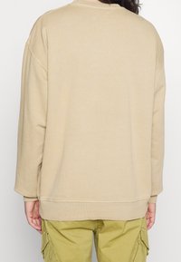 Sudadera beige de gran tamaño con cuello redondo y dobladillo acanalado. Textura suave, mangas largas y aberturas laterales para mayor comodidad. Combinada con pantalones de color verde oliva.