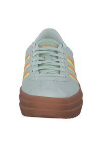 Lichtgrijze suède sneaker met oranje en gele zijstripes, ronde neus, platte bruine rubberen zool en vetersluiting aan de voorkant.