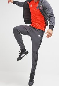 Grijze trainingsoutfit met een oranje shirt, met zwarte en rode accenten. Inclusief zwarte sportschoenen met witte strepen. Materiaal is sportstof.