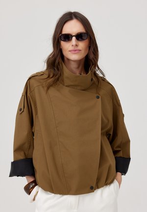 Kobieta nosząca przesadnie dużą brązową kurtkę z czarnymi mankietami, okulary przeciwsłoneczne i białe spodnie na gładkim jasnym tle.