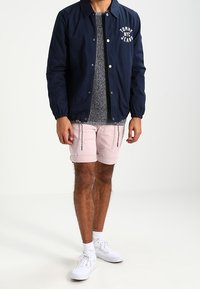 Marinejacke mit elastischen Bündchen und "Tommy NYC Jeans"-Logo, grauer gemusterter Pullover, rosa Shorts mit Reißverschlussdetails, weiße Sneakers.