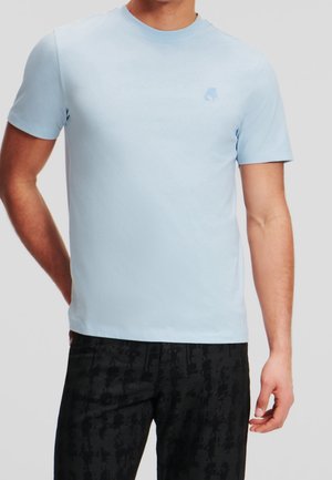 Homme portant un t-shirt bleu clair à manches courtes et un pantalon noir à motifs, debout avec une main derrière le dos.