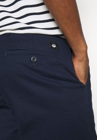 Marinblå byxor i mjukt, texturerat material, med en bakficka som har en svart etikett och en knappstängning. Randig skjorta delvis synlig.