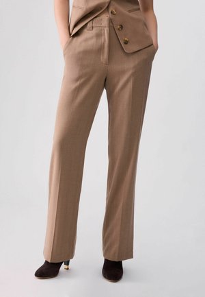 Persona con pantalones beige a medida con bolsillos, combinados con un chaleco beige a juego y zapatos negros de tacón sobre un fondo liso.