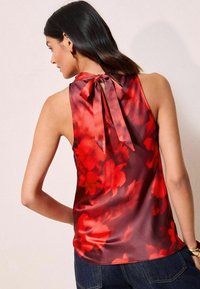 Blusa in satin floreale rossa con un nodo sul collo posteriore, caratterizzata da una texture liscia e design senza maniche. Toni più scuri creano un motivo a gradiente.