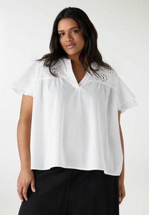 Donna con capelli lunghi e scuri che indossa una camicetta bianca larga con dettagli a occhielli sulle spalle e pantaloni neri su uno sfondo uniforme.