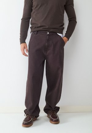Man draagt donkerbruine wijde broekspijpen, een bruine blouse met lange mouwen en donkerbruine veterschoenen, staat met één hand in de zak.
