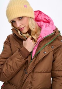 Hnedá nadýchaná bunda s ružovou podšívkou, zelený zips a cvoky. Doplnená svetložltou rebrovanou čiapkou. Mäkká textúra.