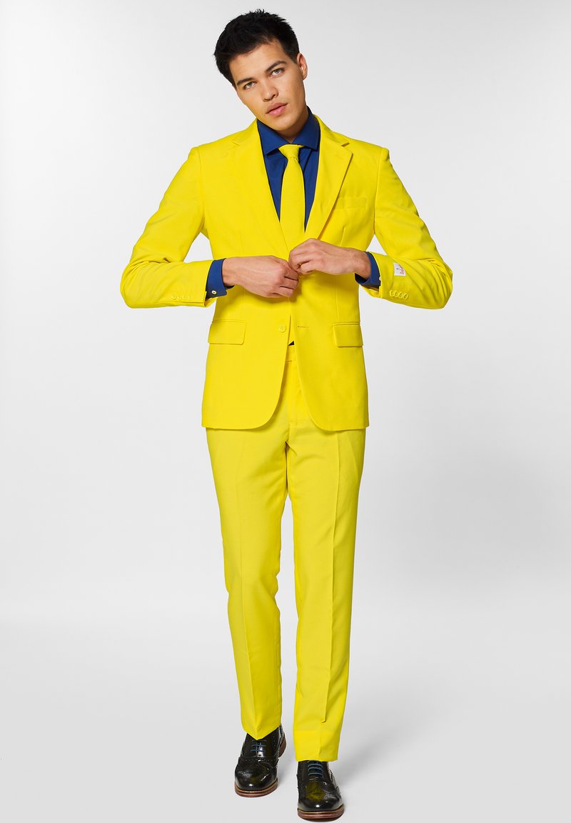 Suit giallo brillante con design su misura, revers strutturati e cravatta abbinata su una camicia blu scuro. Scarpe nere completano l'outfit.
