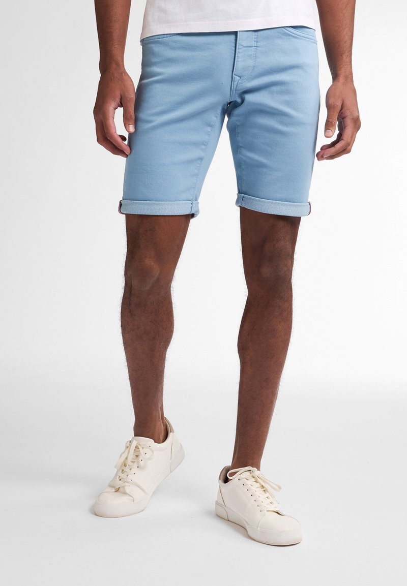 Pantalones cortos de mezclilla azul claro con puños doblados, que cuentan con presillas para cinturón y bolsillos frontales. Combinados con zapatillas off-white.