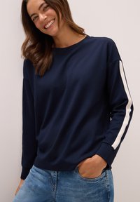 Sweatshirt bleu marine en tissu doux avec un col rond et des rayures blanches sur les côtés ; associé à un jean bleu clair.