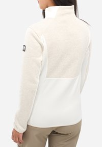 Frau in beiger und weißer Fleecejacke mit hohem Kragen und Millet-Logo am linken Ärmel, steht vor einem schlichten Hintergrund.