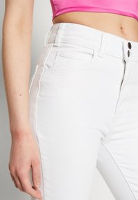 Jeans blancos de cintura alta con cremallera, dos bolsillos frontales y una textura de denim suave y ligera. Costuras visibles en un color a juego.