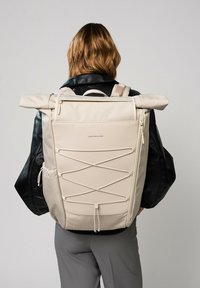 Sac à dos beige avec rabat enroulable, panneaux texturés et détails en cordon élastique. Comprend une poignée supérieure et des fermetures à glissière. Bretelles ajustables.