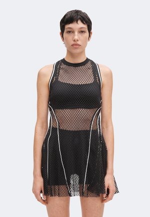 Jong persoon met kort donker haar, die een mouwloze zwarte mesh-jurk met witte bies draagt over een zwarte sportbeha en rok.