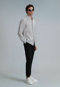Gestreepte langemouwen shirt in beige en bruin, aangesloten zwarte broek en witte sneakers. Model draagt donkere zonnebril en poseert tegen een grijze achtergrond.