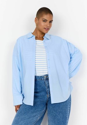 Persoană cu păr scurt, purtând o cămașă deschisă la culoare peste un tricou în dungi albe și albastre și blugi albaștri cu talie înaltă, pozând.