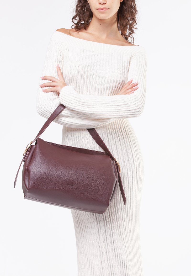 Bordeaux lædertaske med rund form, kort brun rem og subtil tekstur. Har et minimalistisk design og en lille logo detalje.