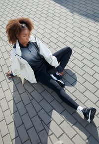 Tenue de sport composée d'une veste blanche, d'un t-shirt à manches longues gris foncé, de leggings noirs et de baskets noires avec des accents blancs, sur un sol pavé.