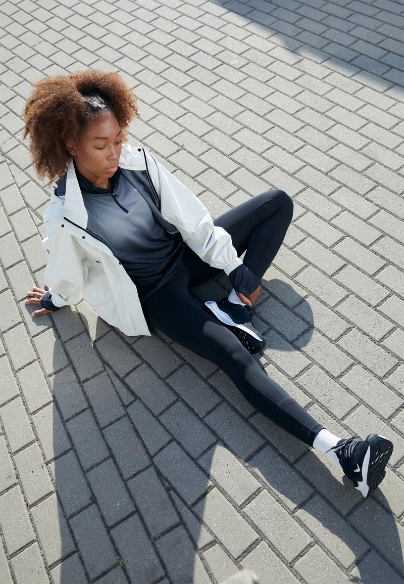 Sportowy strój składający się z białej kurtki, ciemnoszarej koszulki z długim rękawem, czarnych legginsów i czarnych sneakersów z białymi akcentami, na utwardzonej nawierzchni.