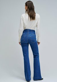 Vaqueros de tiro alto y acampanados en denim azul oscuro con tejido texturizado, que presentan bolsillos traseros y detalles de costura en contraste.