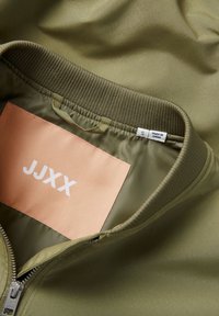 Gros plan sur le col d'une veste vert olive avec une étiquette couleur pêche indiquant "JJXX" et une étiquette blanche de taille marquée "S Made in China.".