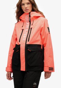 Jasnopomarańczowa i czarna wodoodporna kurtka z kapturem, z dwoma przednimi kieszeniami, odblaskowym logo i regulowanymi mankietami.