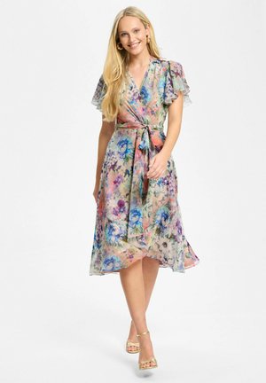 Smilende kvinde går iført en midlængde blomstret slå-om-kjole med korte flagrende ærmer og guldfarvede hæle med ankelrem på hvid baggrund.