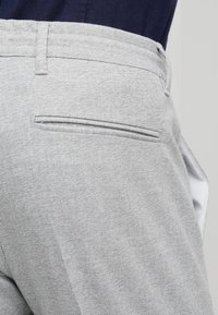 Šedé kalhoty s texturovaným tkaným vzorem, které mají jednu zadní kapsu. Pas má hladký povrch a poutka na opasek.