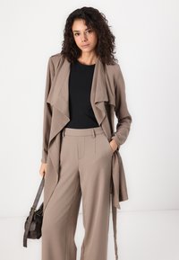 Lys brun draperet cardigan over en sort top, parret med matchende højtaljede bredbenede bukser. Brun håndtaske fremhæver outfittet.