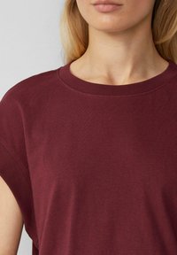 Burgunderfarbenes kurzärmeliges Baumwolloberteil mit rundem Ausschnitt. Es hat eine glatte, weiche Textur und einen lockeren Schnitt, der den Schulterbereich betont.