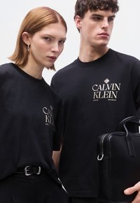 Μαύρα T-shirt με λογότυπο και κείμενο σε κρεμ χρώμα και κλασική γραμματοσειρά. Ένα άτομο κρατά μια μαύρη δερμάτινη τσάντα με ορθογώνιο σχήμα και διπλές λαβές.