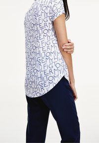 Camisa blanca de manga corta con un patrón de texto en color marino, con un dobladillo redondeado y aberturas laterales. Combinada con pantalones marinos, mostrando un ajuste relajado.