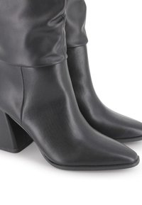 Botas de cuero negras hasta el tobillo con puntas afiladas y tacones cuadrados, mostradas sobre un fondo blanco.