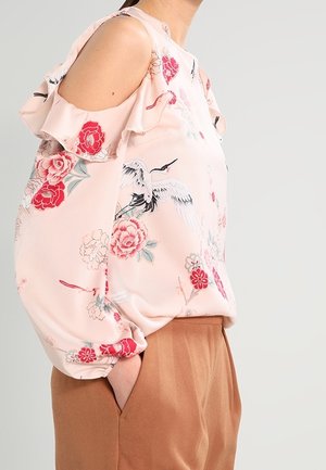Femme portant un chemisier rose à épaules dénudées avec imprimé grue et fleurs, associé à un pantalon marron taille haute, mains dans les poches.