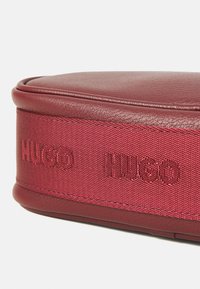 Rund rød lærveske med rød stoffbånd sydd fast, preget med "HUGO"-logo i matchende tråd.