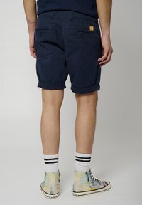 Pantalones cortos azul marino con puños enrollados, dos bolsillos traseros con botones y cremalleras, y un parche de logo naranja. El modelo lleva zapatillas altas con estampado.