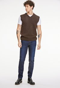 Brun ärmlös stickad tröja med V-ringning över en vit T-shirt, kombinerad med mörkblå smala jeans och svarta ankelboots.