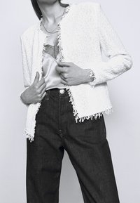 Biała teksturowana kurtka z postrzępionymi krawędziami, zestawiona z satynowym topem i ciemnymi dżinsami. Tkanina ma wzór warkoczowy.